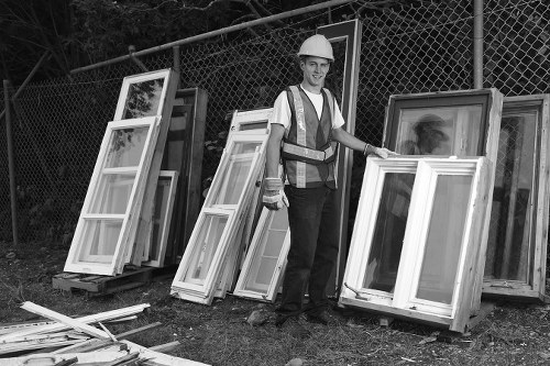 A male worker wearing a safety helmet and high-visibility vest stands outdoors on a grassy area near a chain-link fence, surrounded by several large, white, double-glazed window frames leaning against the fence and placed on wooden pallets. The window frames feature white PVC or timber finishes with glass panes and are positioned in a slight angled arrangement, some leaning more upright while others are tilted. In front of the frames, a few loose wooden or PVC sash components lie on the ground. The scene appears to be part of a waste removal or clearance operation, consistent with a rubbish disposal service, possibly in a suburban or industrial area near Alperton. The environment is lit naturally, and the overall setting is organized for collection or transportation of waste materials related to garden or property clearance. The image visually supports services provided by Garden Clearance Alperton in waste management and rubbish removal tasks involving window frames and similar materials.