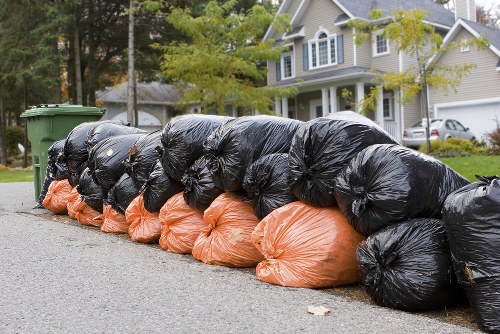 A row of large black plastic rubbish bags and orange refuse sacks stacked along the curbside of a residential street, likely for collection or disposal. The black bags are tightly packed and appear to contain garden waste, household rubbish, or general refuse, with some slightly bulging and tied at the top. The orange sacks are visibly filled and placed underneath the black bags, which are arranged in a neat line along the pavement during daylight hours. In the background, there are green wheelie bins, a paved road, and a row of modern houses with white and beige facades, gabled roofs, and well-maintained front gardens. Trees with green leaves partially shade the scene, indicating a pleasant, suburban environment. The overall setting suggests ongoing or completed rubbish removal activity in an area serviced by Garden Clearance Alperton, located within the London boroughs, and highlights typical urban waste collection practices for household or garden waste management.