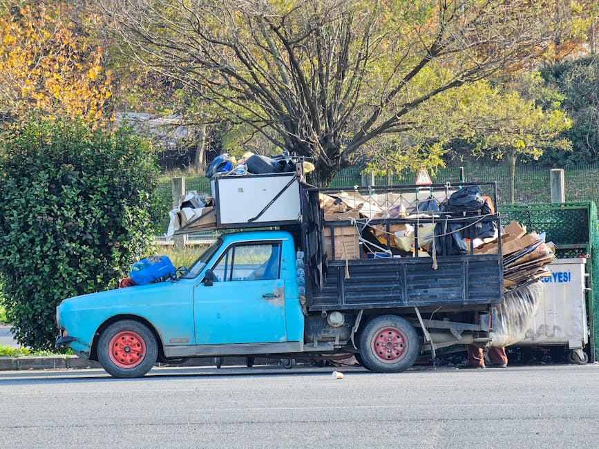 A small burgundy flatbed truck equipped with a protective metal frame on the cab's rear section, parked on an urban street in front of a modern building with large glass windows. The truck's cargo bed