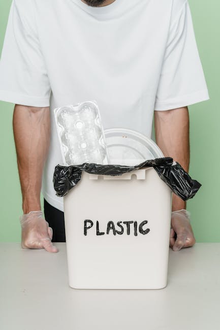 A close-up view of a person's hand placing a crumpled brown paper bag into a grey rubbish bin with a white lid, situated indoors on a wooden surface. The person's arm is partially visible, wearing a w