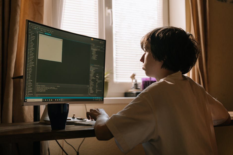 A person with blond hair, wearing a white shirt, is seated at a desk in an industrial-style office or workspace. They are facing away from the camera, working on a large desktop computer displaying li